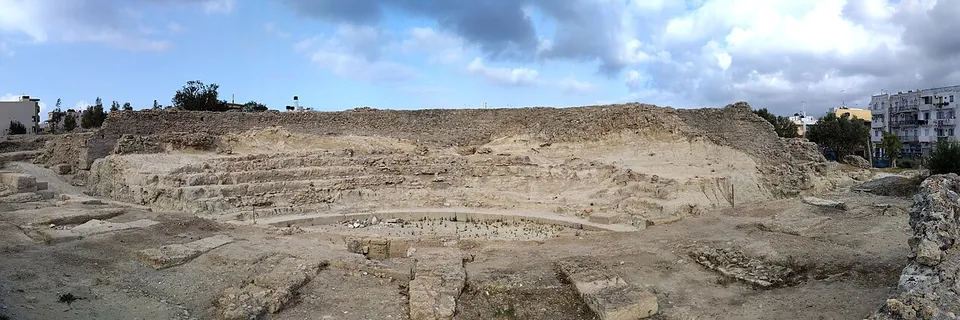 A wide view of an excavated theatre cut into a low hill of pale earth, with rows of seats around the orchestra and blocks of the stage in the foreground.