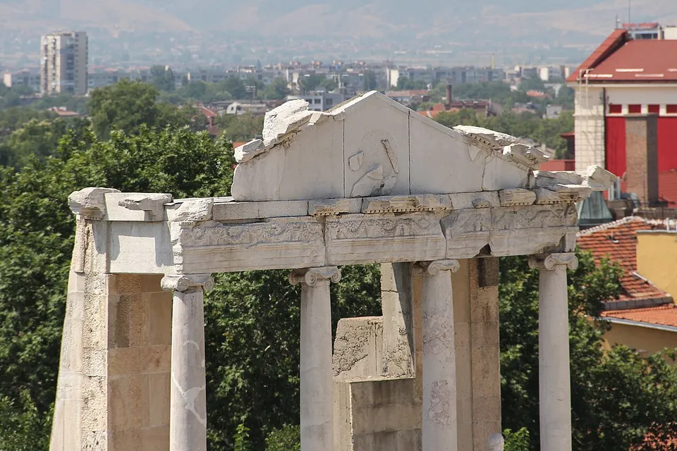 A broken marble pediment with a worn relief at its centre, resting on a carved frieze and four white columns with scrolled capitals, against trees, red roofs and tower blocks.