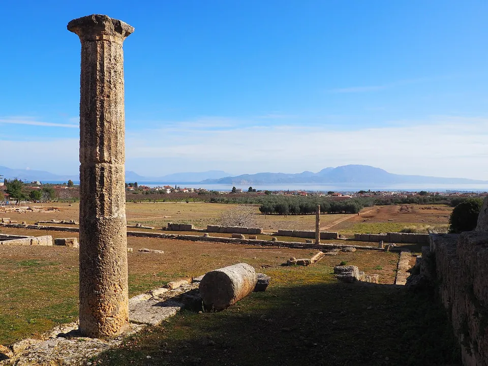 A single weathered stone column standing beside a fallen drum on a grassy plateau, with low walls of the excavated city, the plain and the sea and mountains beyond.