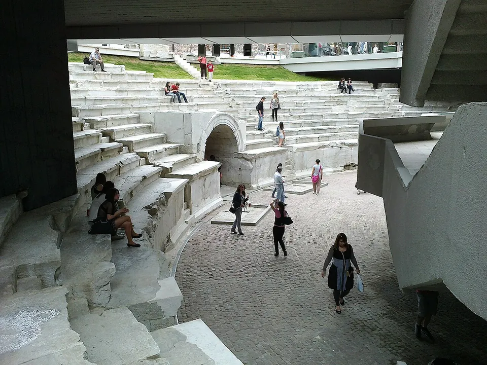 The sunken curved end of the stadium seen from beneath a modern concrete overhang, its marble tiers and arched entrance below a lawn and the street level, with people walking across the paved floor and sitting on the seats.