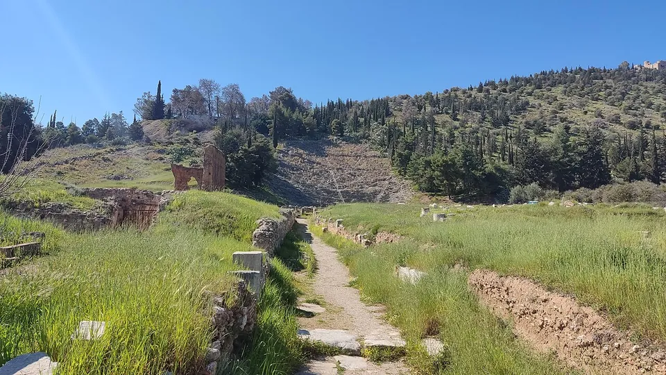 A stone path through long green grass towards a steep hillside with the rock-cut rows of a great theatre, a brick ruin on the left and a castle on the hilltop.
