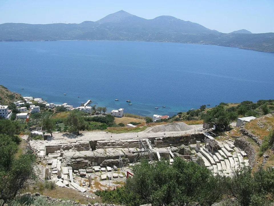 Looking down on a ruined stage building of grey stone and marble blocks and the lower rows of a marble theatre, with the white houses of a small harbour and a broad bay beyond.
