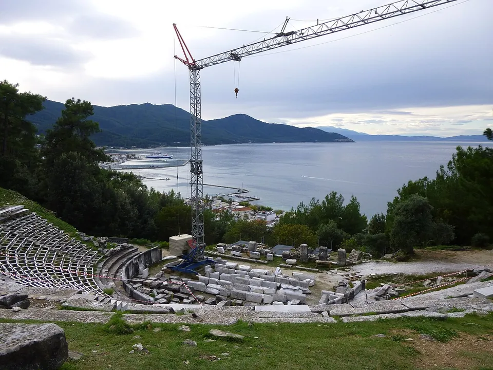 The curved stone seating of a theatre on a wooded slope, with a construction crane, marble blocks laid out on the ground and the town and a wide bay with mountains beyond.