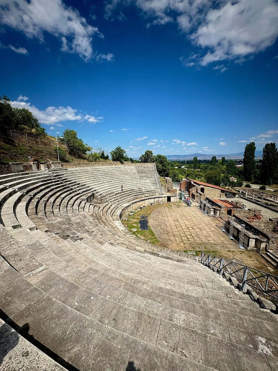 Curving rows of pale stone seats sweeping down to a paved semicircular orchestra, seen from the top of the theatre, with brick and stone ruins beside the stage, trees and distant mountains under a blue sky.