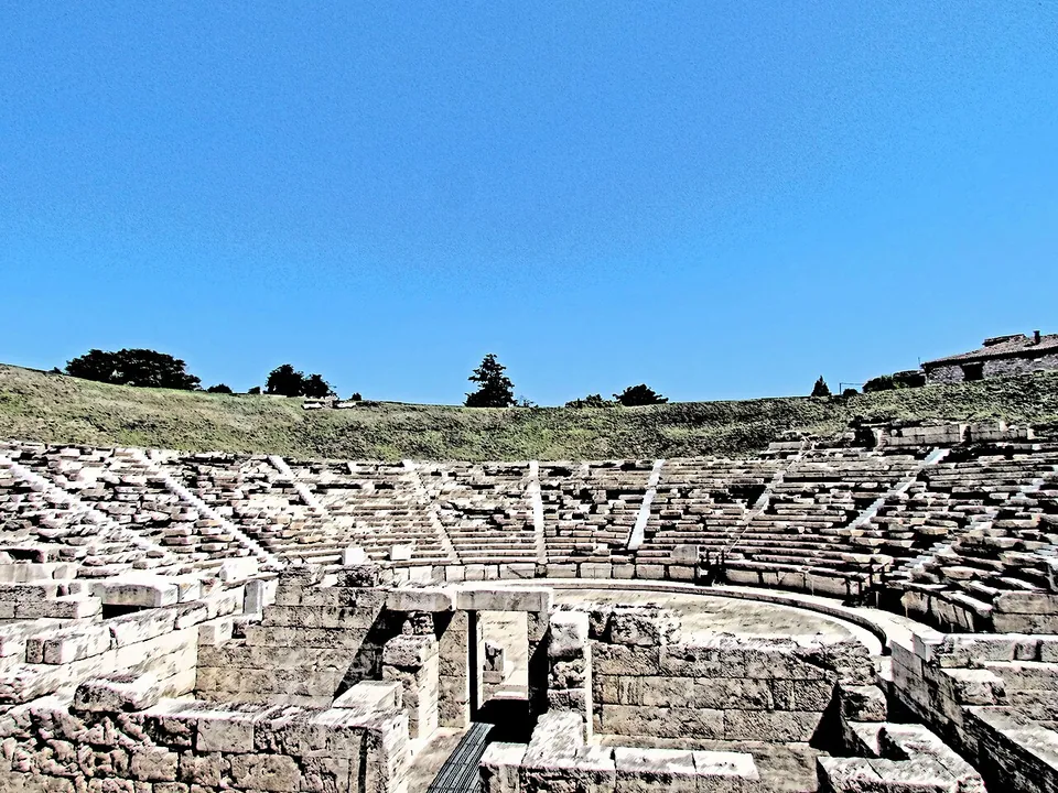 Rows of pale marble seats climbing a grassy hillside in a wide semicircle, divided by stairways, above the stone walls of the stage building in the foreground.