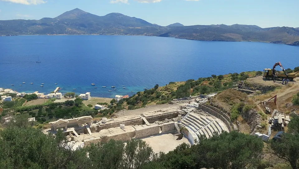 A small white marble theatre with curved rows of seats and a long stage building, set on a scrubby hillside above a wide blue bay with boats, a few white houses at the shore and mountains across the water.
