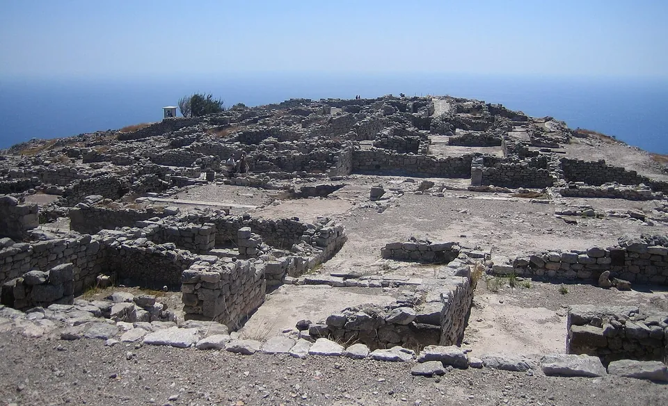 A long ridge-top city of low grey stone walls, house plots and paved lanes under a hazy sky, with the sea on both sides.