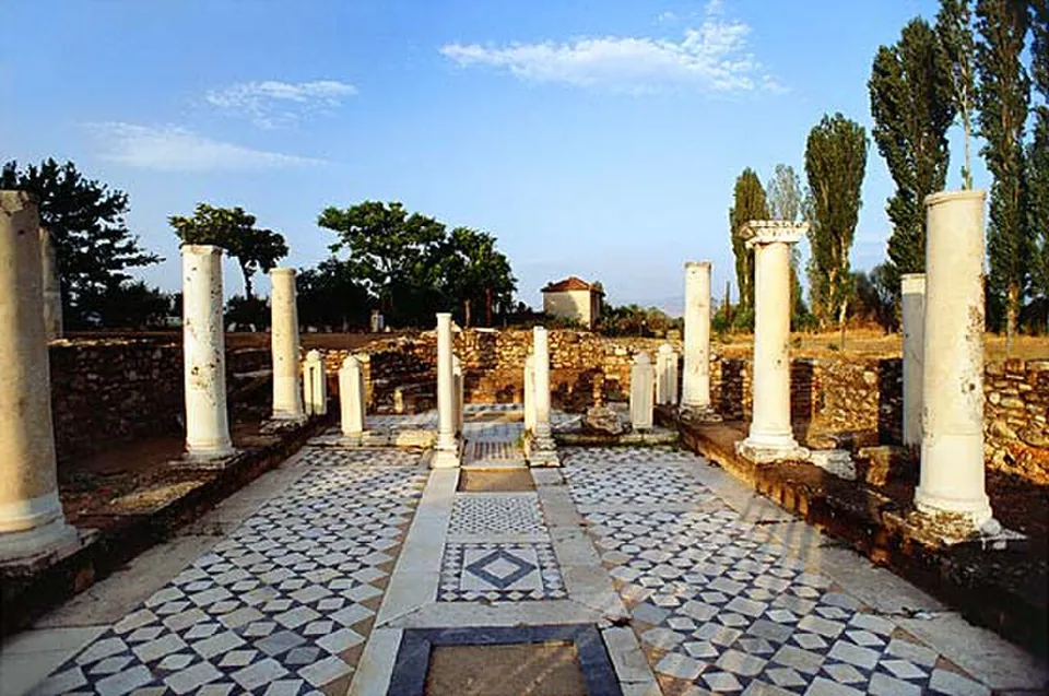 A colonnaded court with white marble columns, some broken off, standing along a floor of black and white geometric mosaic, with low stone walls, poplars and trees behind.