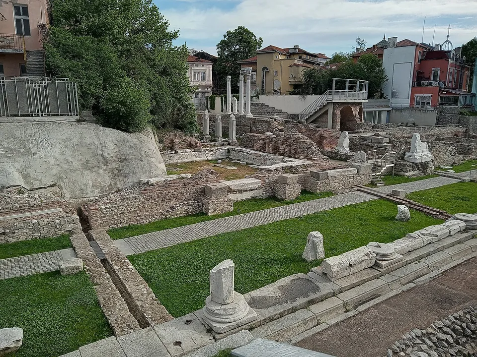 Excavated stone walls, paved strips and column bases in mown grass, with a few re-erected white columns and the houses of the city centre behind.