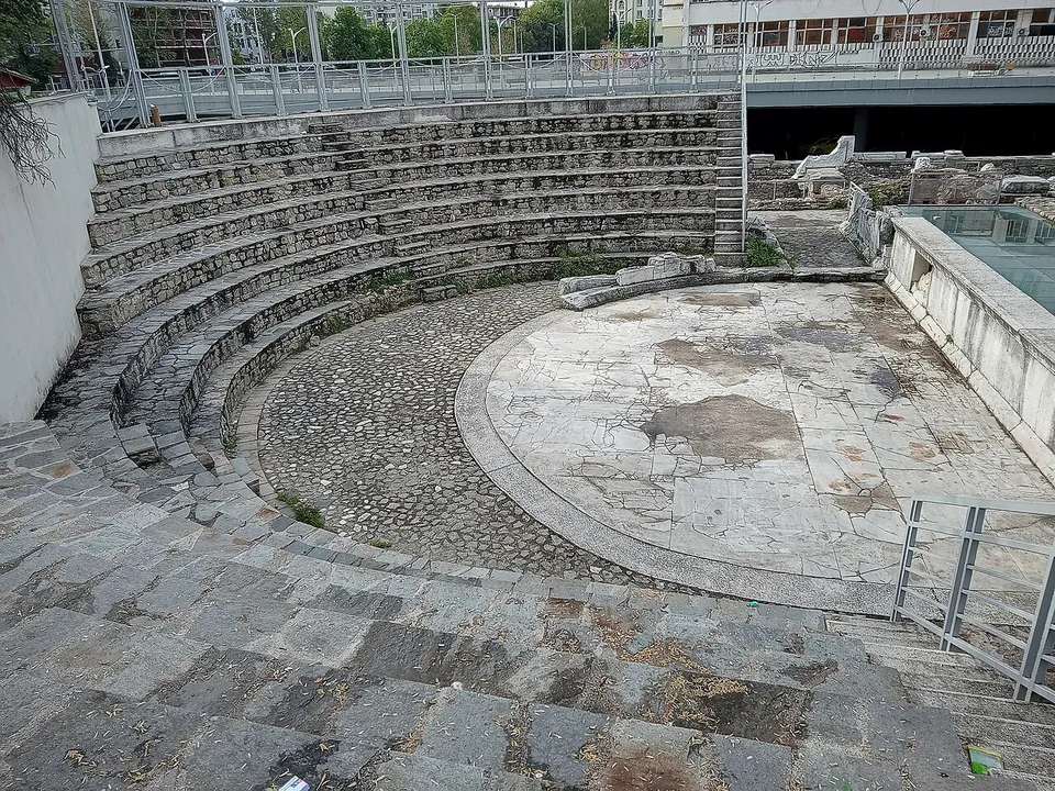 A small semicircular auditorium of stone seats rising in steps round a paved orchestra floor of large worn marble slabs, with a metal railing and a road bridge above the top row.