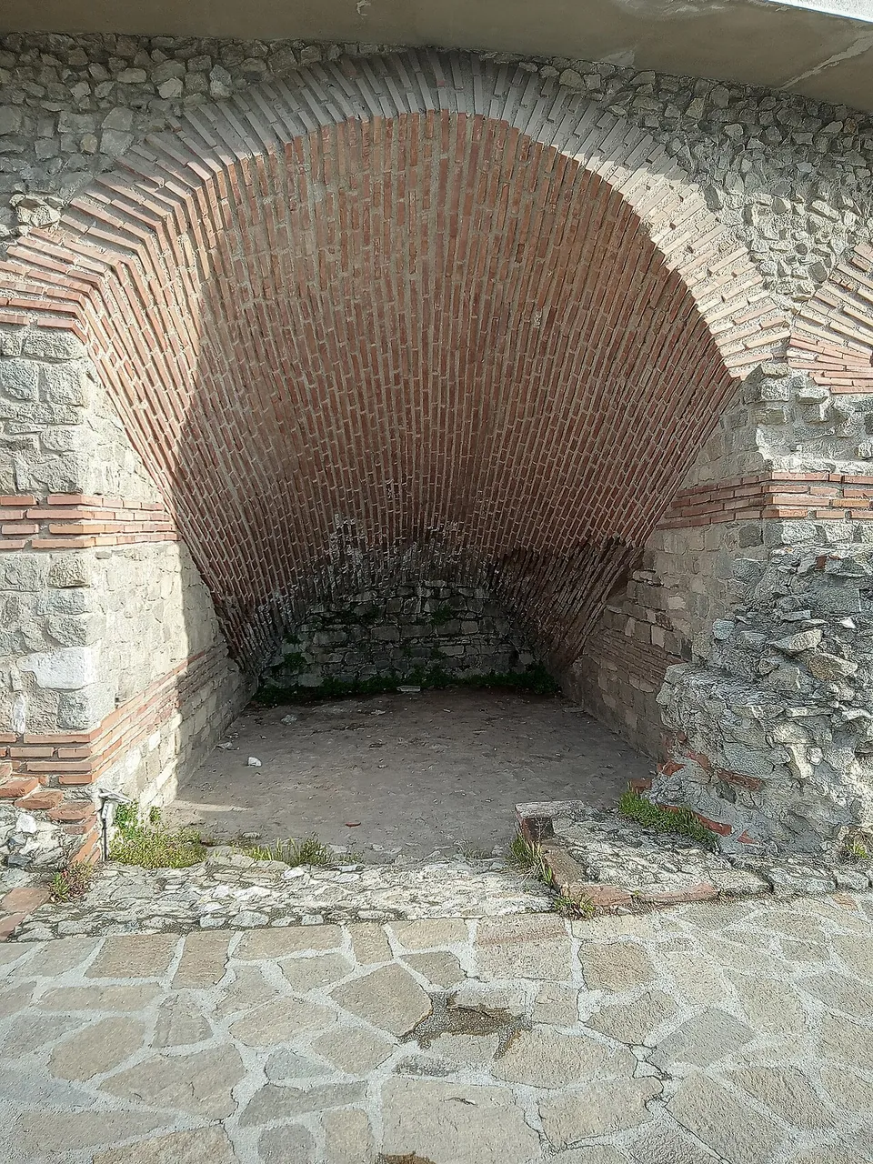 A deep vault of red brick laid in a fan pattern, springing from walls of rough stone banded with brick, opening onto a paved path.