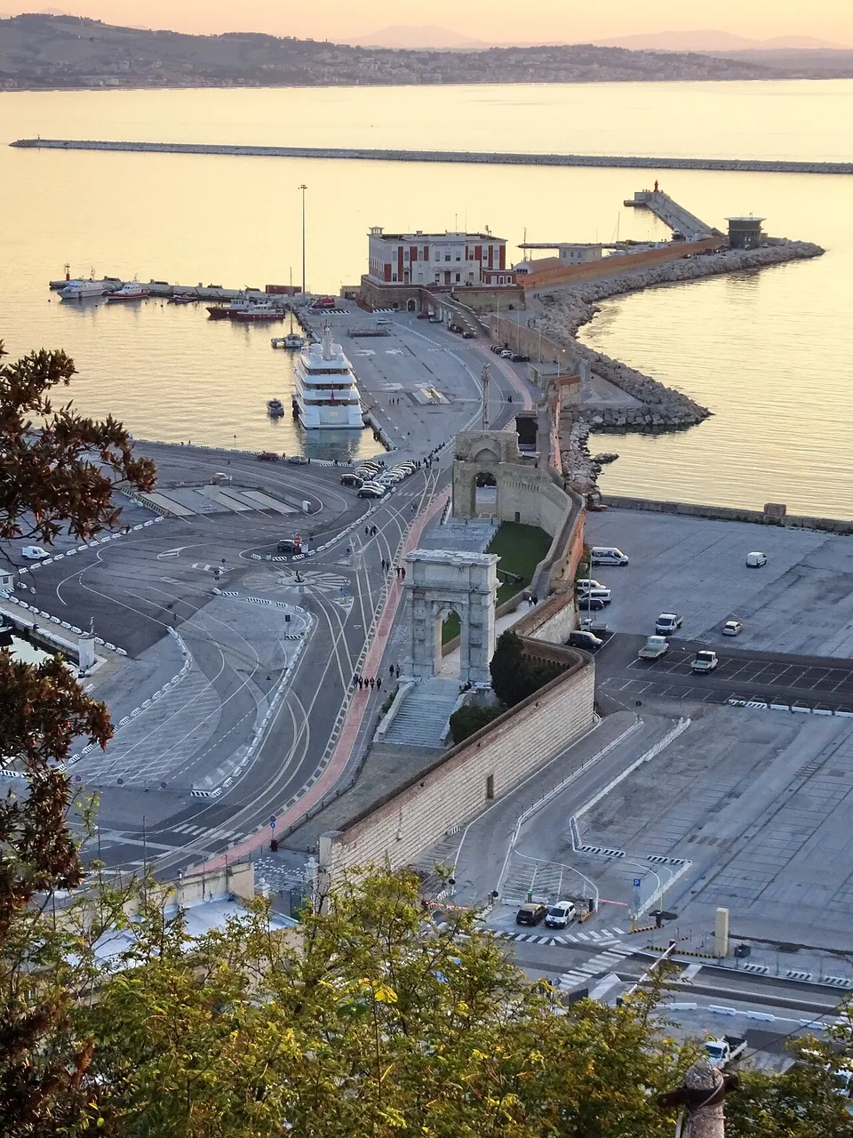 The port of Ancona seen from the hill above at sunset, a long quay running out between wide paved docks and parked cars, with a small white marble arch standing on it and a second arch farther along, a breakwater and calm sea beyond.