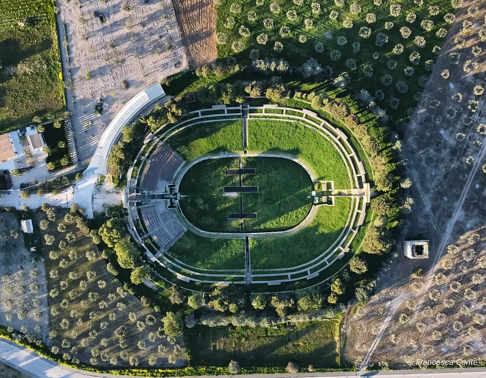 A drone view looking straight down on the amphitheatre, an oval of grass ringed by low walls and tiers of seating, with dark lines crossing the arena floor, set among olive groves beside a road.