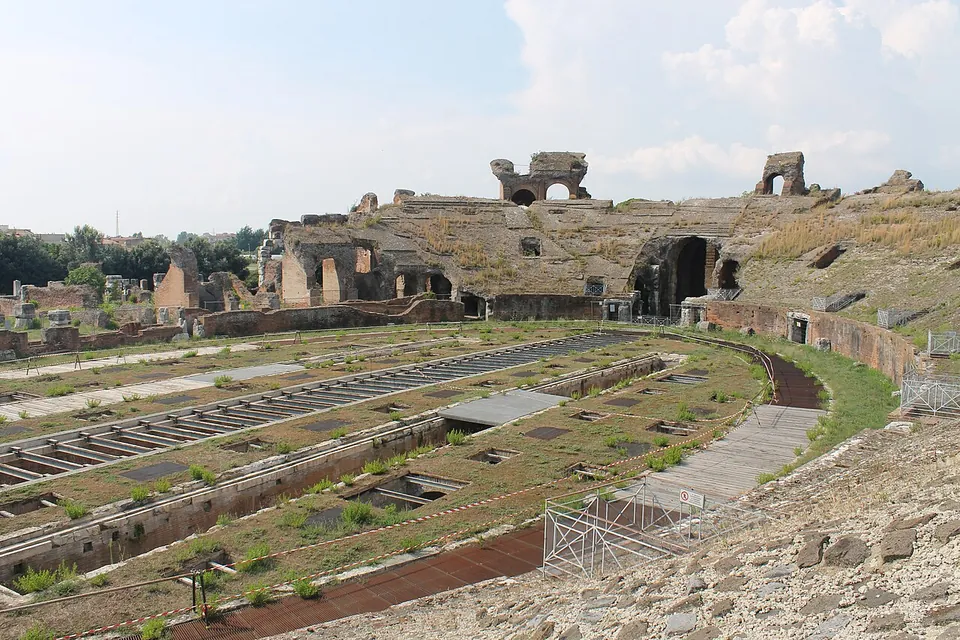 Seen from the ruined seating, the long arena floor with rows of openings and grilles over the passages beneath it, ringed by the broken mass of the cavea, with two arches standing high on its rim against the sky.