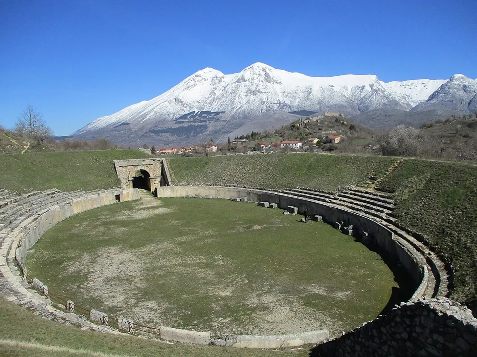 An oval arena of mown grass ringed by a stone parapet and grassy banks of seating, with an arched gateway at the far end and a snow-covered mountain range under a clear blue sky behind.