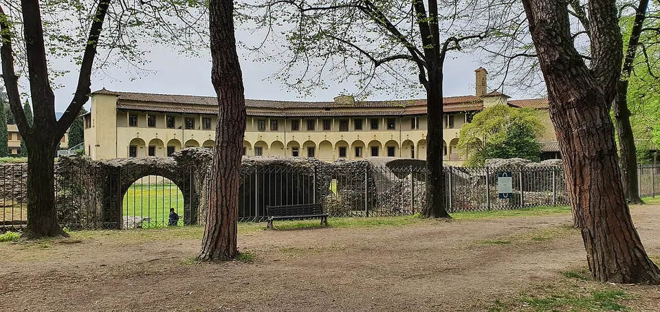 A shaded path under tall trees beside a low ruined wall pierced by a round arch, with a fence along it, a bench, and the arcaded museum building beyond.