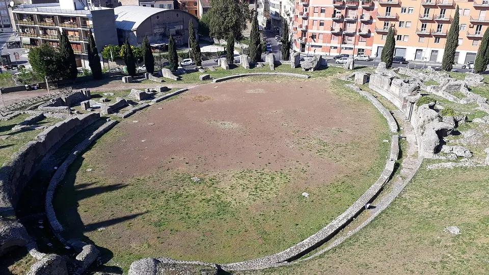 The oval arena of an amphitheatre seen from above, a grassy floor ringed by low stone walls and scattered blocks, with cypresses and modern apartment blocks around it.