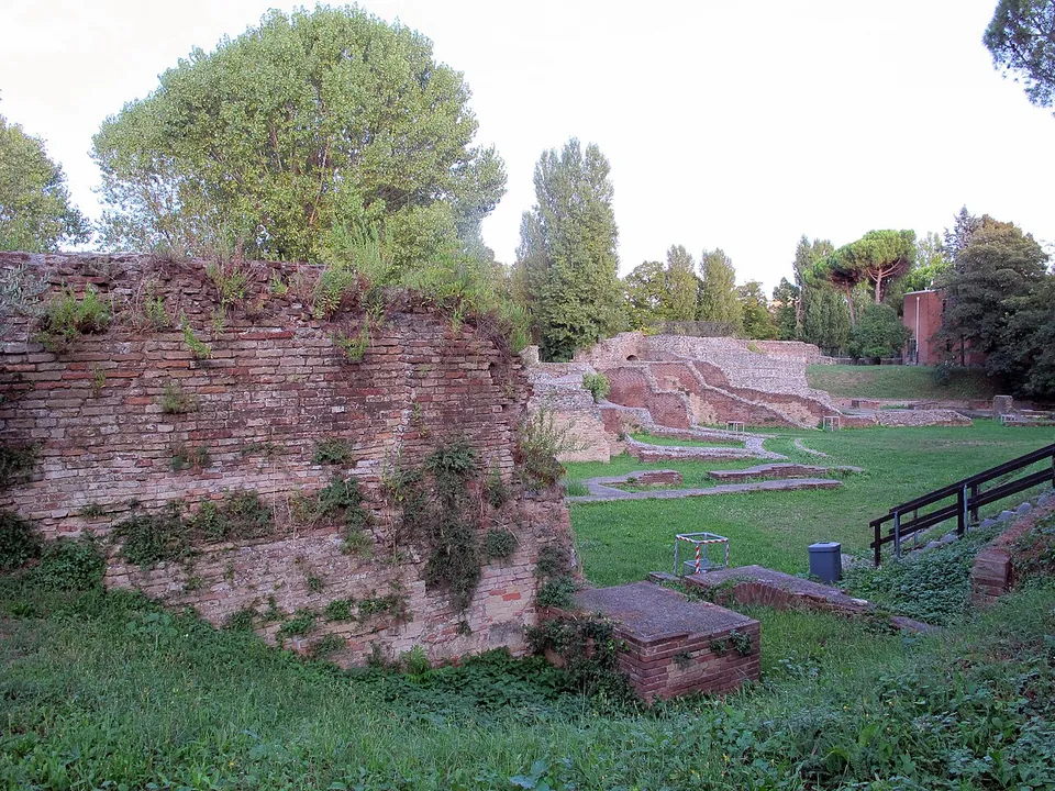 A thick wall of red brick overgrown with plants in the foreground; beyond it a lawn with the low outlines of excavated walls, a railing, and at the back a stepped bank of pale masonry under trees.