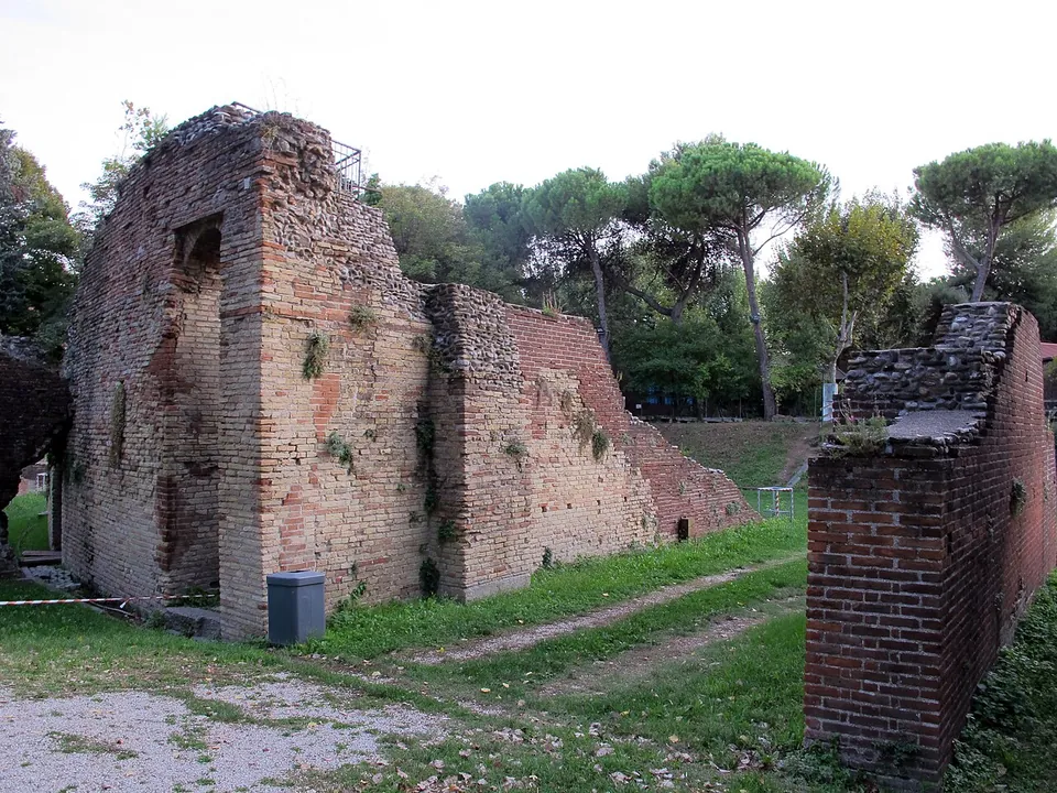 A tall block of brick masonry with a dark doorway-like opening and a lower stretch of wall beside it, on a grassy path under umbrella pines; a separate brick fragment stands at the right.
