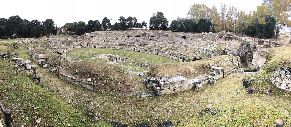 A wide view across the grassy oval of the amphitheatre, ringed by low walls and rock-cut tiers, with a sunken rectangular pit in the middle of the arena and a line of trees beyond.