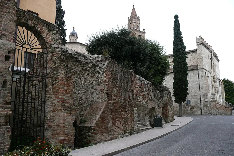 A long curving wall of brick and rubble lining a street pavement, with a brick arch closed by an iron gate at its near end, and the bell tower and stone flank of the cathedral rising behind trees at the far end.