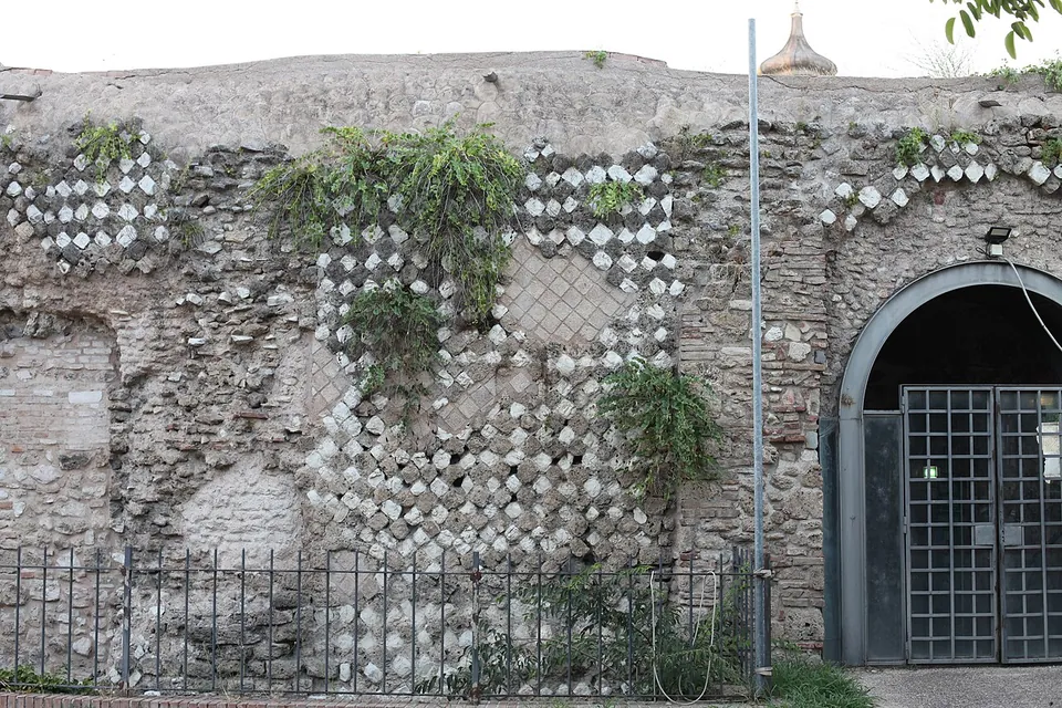 The outer face of the amphitheatre behind a low railing, its patched stonework showing the outlines of closed arches and diamond-pattern facing, with a modern iron gate set in an arch at the right.