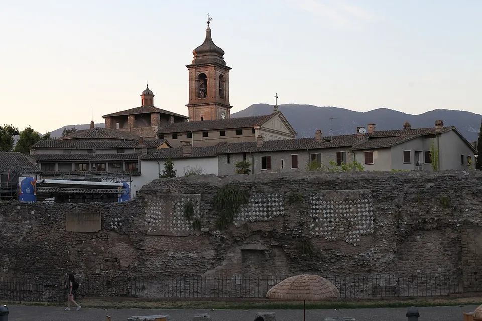 A long, low stretch of ancient wall along a pavement, patched with panels of diamond-pattern stonework, with the roofs of the old town and a bell tower rising behind it and mountains on the horizon.