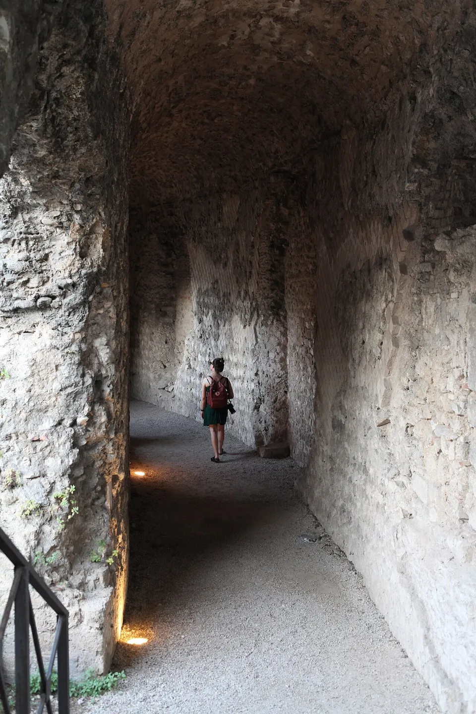 A long, curving barrel-vaulted corridor of rough stone, lit by small lamps along the floor, with one visitor walking away down it.