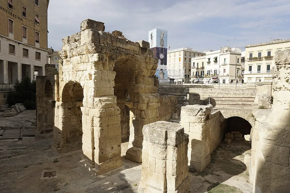 A block of massive squared stones pierced by two round arches, with a lone stone pier in front, a low arched passage in a wall to the right, and apartment buildings above under a blue sky.