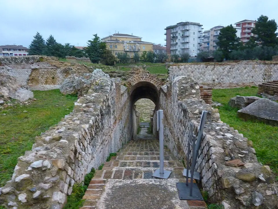 A narrow arched passage between rough stone walls, its sloping floor paved with brick, with a wall of net-pattern stonework behind and apartment blocks rising over the trees.