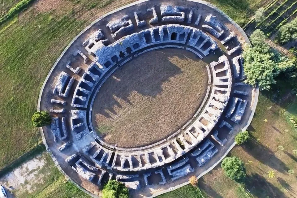 An aerial view of an oval amphitheatre, its arena of bare earth ringed by the stubs of stone walls that divide the seating into rows of small chambers, set in mown grass with trees around it and a path running past one end.