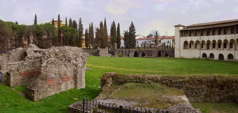 Rough masses of Roman masonry with bands of red brick rising from the grass in the foreground, the open lawn of the arena beyond, and the arcaded museum building and a line of cypresses behind.