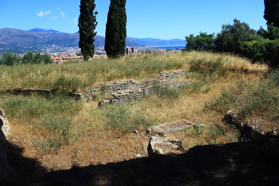 A dry grassy hollow edged by low courses of stone wall, with cypresses, the town and the coastal plain spread out beyond.