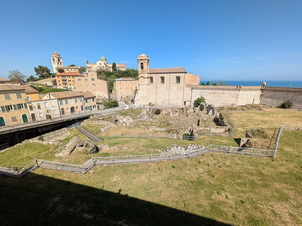 The excavated amphitheatre on a grassy slope, low walls and scattered blocks behind modern railings, with a walkway along one side, a church with a bell tower and houses above, and the sea on the horizon.