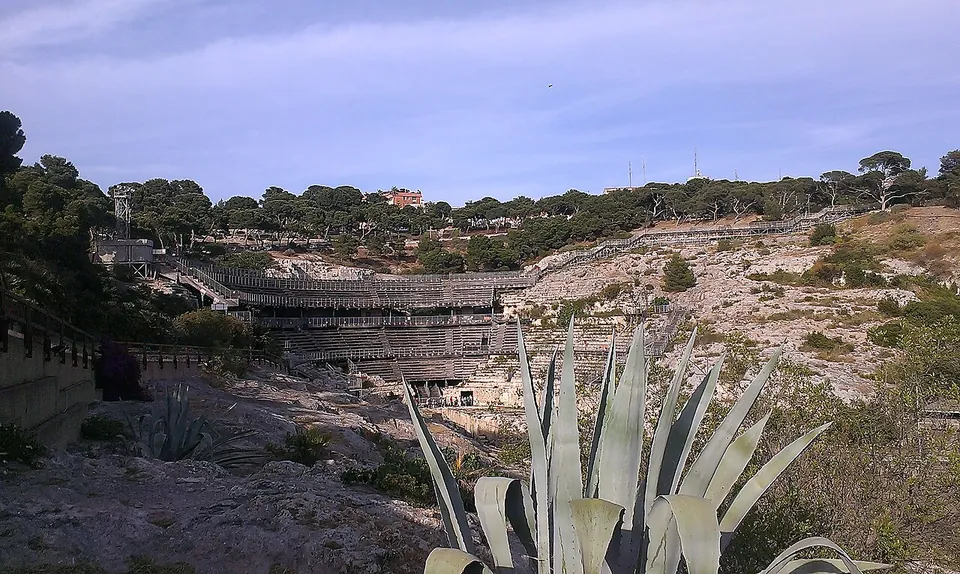 The amphitheatre seen from a distance across a rocky slope with an agave in the foreground, its tiers of seating and a walkway set into a pale limestone hollow under a tree-lined ridge.