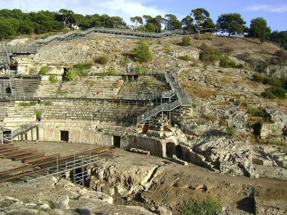 Rows of seating cut into a sloping face of bare limestone, with metal stairways climbing it, and below them the low wall round the arena pierced by rectangular openings.