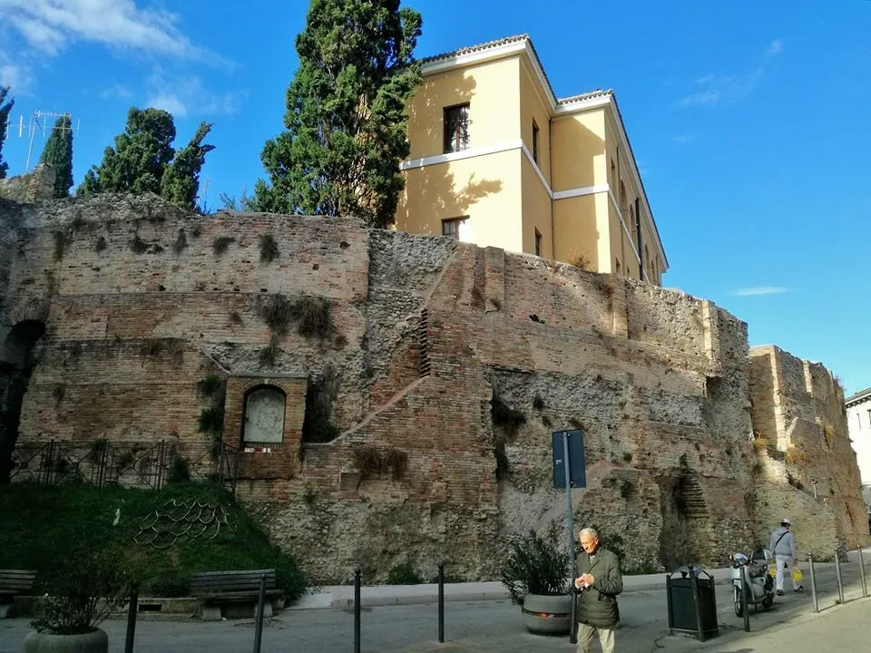 A long, high stretch of weathered brick and stone wall running beside a paved street, with a small arched window set in a niche, trees and a pale yellow building above it, and benches, a street sign and a passer-by in front.