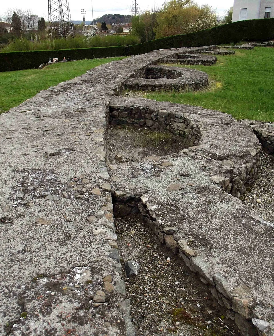 The low stone foundations of the amphitheatre curving away across a mown lawn, a broad band of rubble masonry with a row of semicircular compartments set into it, a hedge, pylons and a building behind.