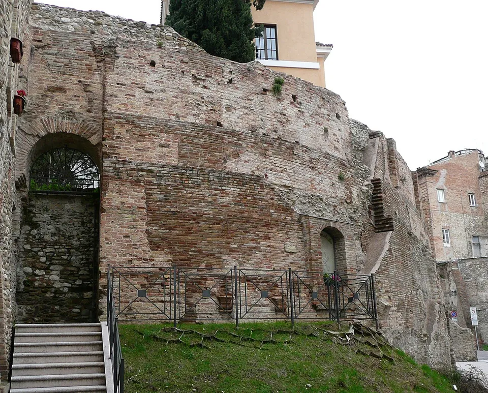 A tall curving wall of red brick patched with grey stone, with a round-headed arch filled by a fan-shaped iron grille and reached by a short flight of steps at its left end, a small arched niche further along, a sloping lawn and railings at its foot, and a pale building above it.