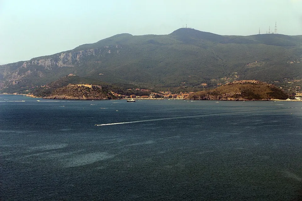 A wide expanse of dark blue sea with a boat's wake, and across the water a small coastal town at the foot of a high green mountain.