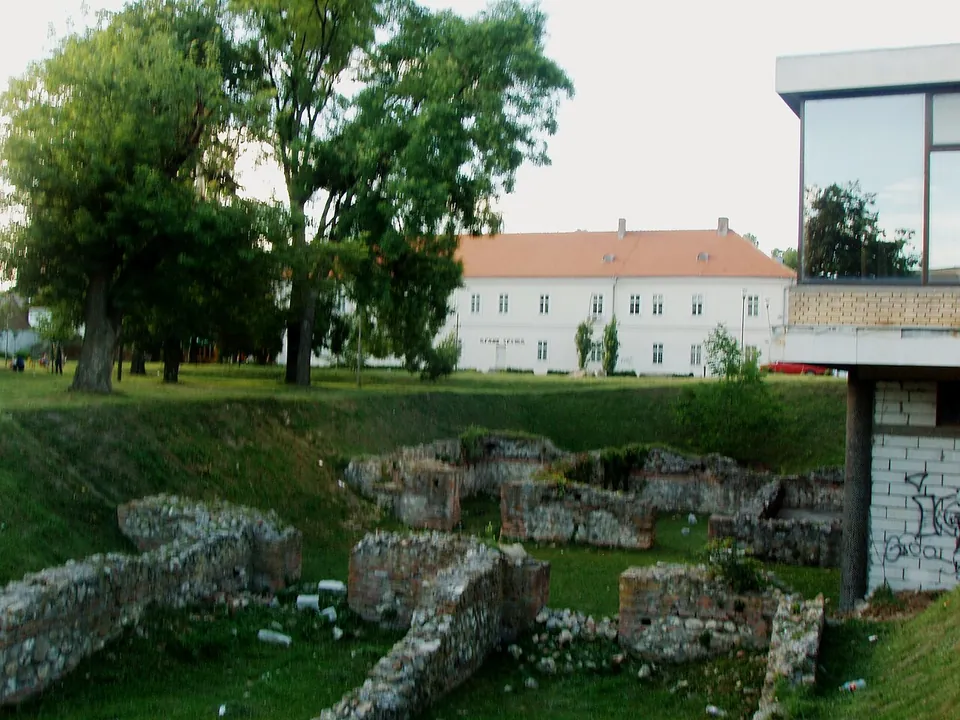 Low walls of rough stone and brick stand in a grassy hollow under trees, with a white two-storey building behind and the corner of a modern glass-fronted building at the right.