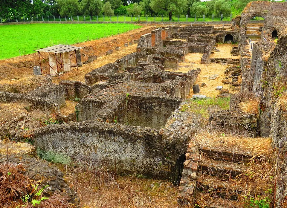 A cluster of excavated rooms with walls of small square stones laid in a net pattern, one curved, standing about a metre high among dry grass, with a temporary shelter and a fenced field behind.