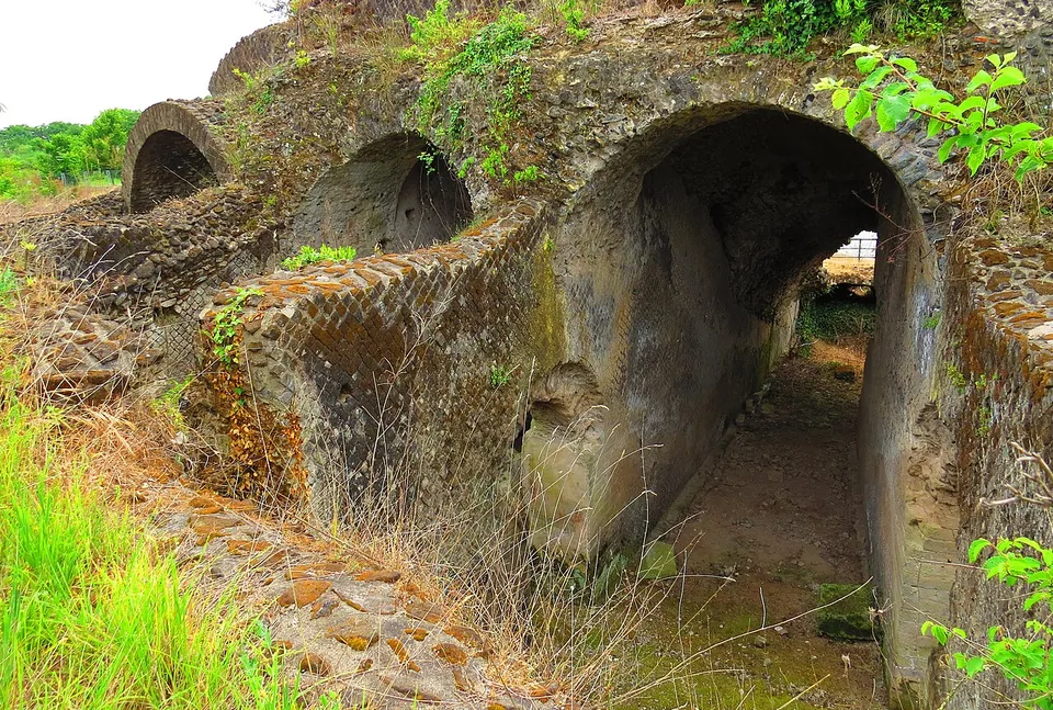 A row of barrel-vaulted passages built of rubble and net-patterned stonework, one opening into a long dark corridor, with grass and shrubs growing over the top.