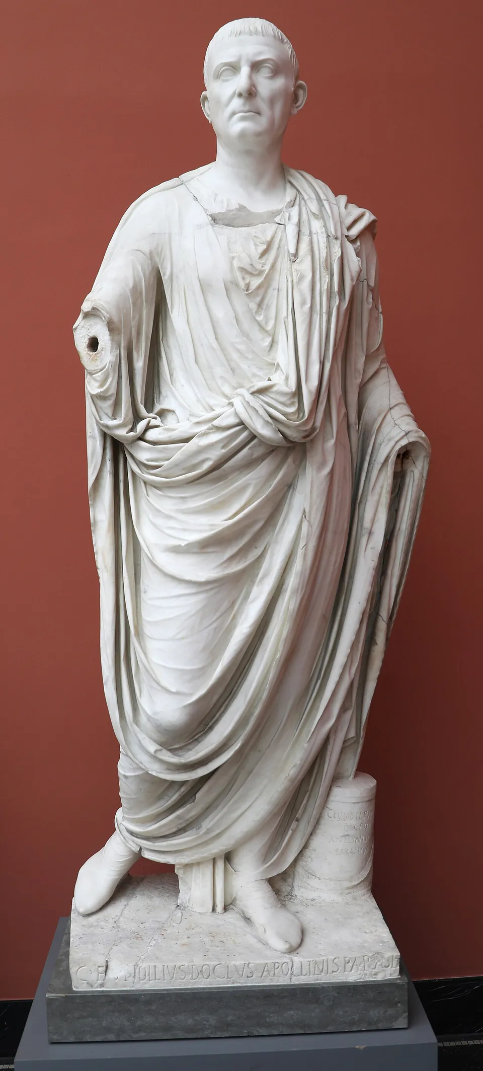A marble statue of a man in a toga, standing on a low base against a dark red museum wall.