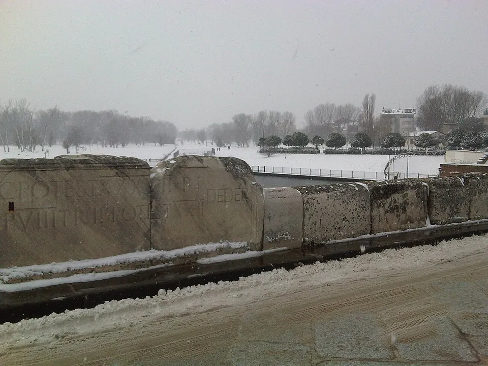 Large stone slabs forming the parapet of the bridge, the nearest two cut with lines of Roman capital letters, with snow on the roadway and a snow-covered park and bare trees beyond.