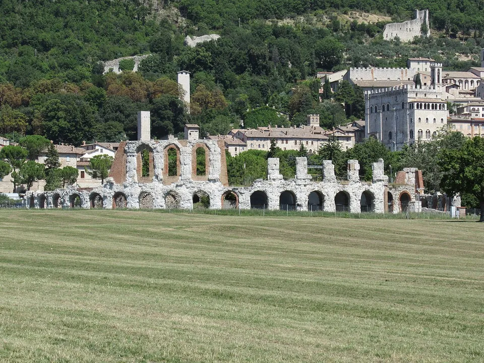 The outer curve of the theatre seen across a mown field - a row of round-headed limestone arches with a taller two-storey stretch of arcade above them - and behind it the stone houses, towers and palaces of the town climbing the hillside.