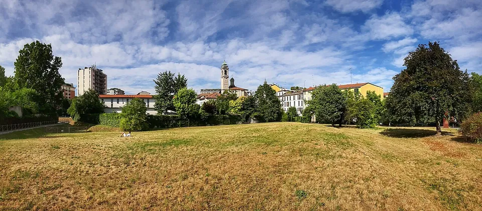 A wide, gently uneven lawn of dry summer grass ringed by trees, with apartment buildings and a church bell tower on the skyline.
