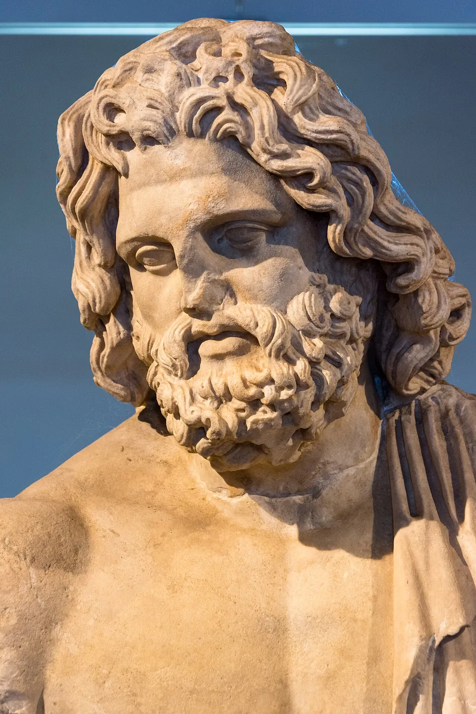 The head and shoulders of a marble statue of a bearded god with thick curling hair and a cloak over one shoulder, in front of a pale blue wall.