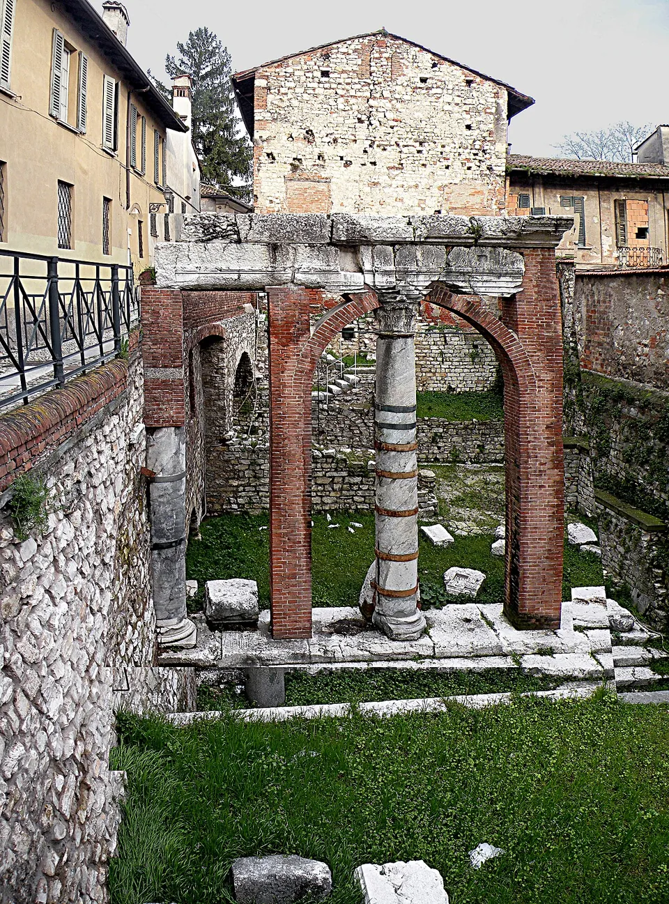 A walled excavation below street level, with a railing along its upper edge, in which a single veined marble column stands between two brick arches that carry a stone beam, on grass scattered with stone blocks.