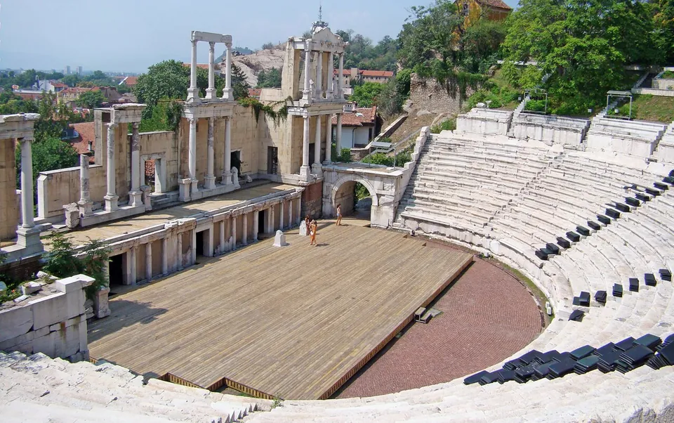 The stage building of the theatre, a two-storey front of white marble columns and pediments with three doorways, behind a wooden stage floor, with the curved rows of marble seats rising on the right.